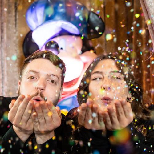 Two UMKC students joyfully blow glitter out of their cupped hands while standing in front of a statue of Mickey Mouse at the Disney 100 exhibit at Union Station